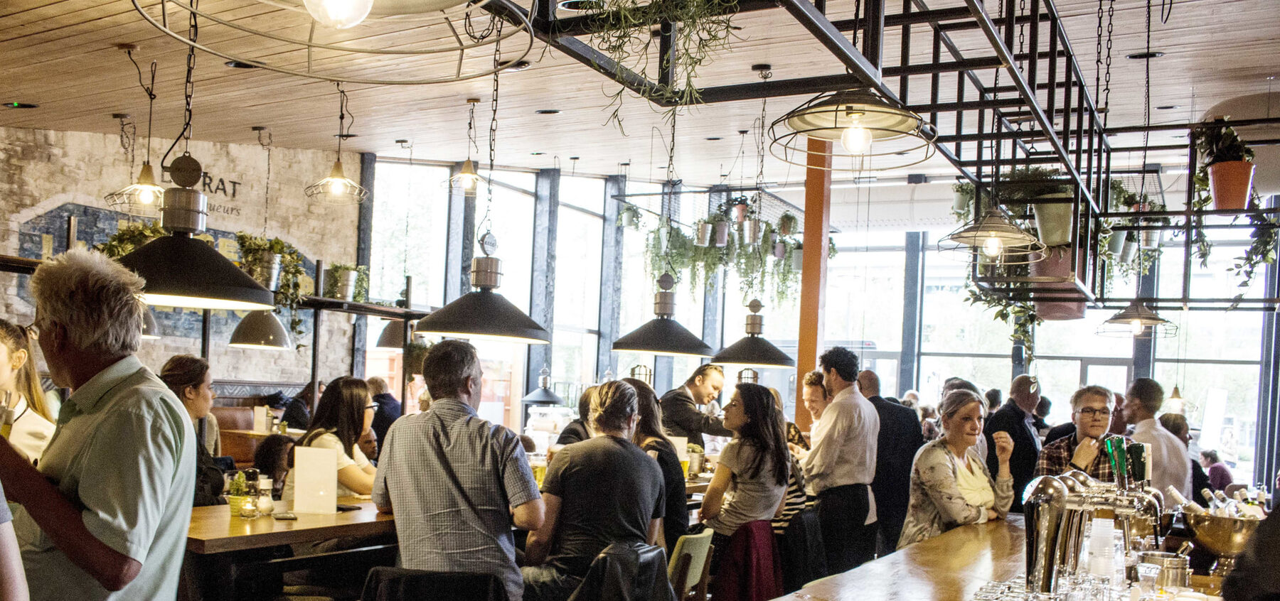 Een volle zaal bij Helden met grote ramen en hanglampen